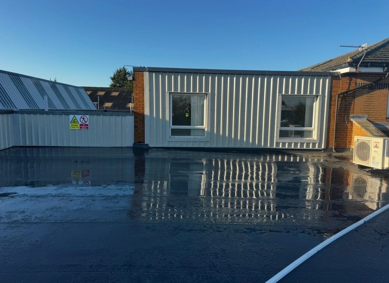 Completed commercial flat roof beside profiled cladding and a plant unit, water sheeting cleanly off the finished membrane