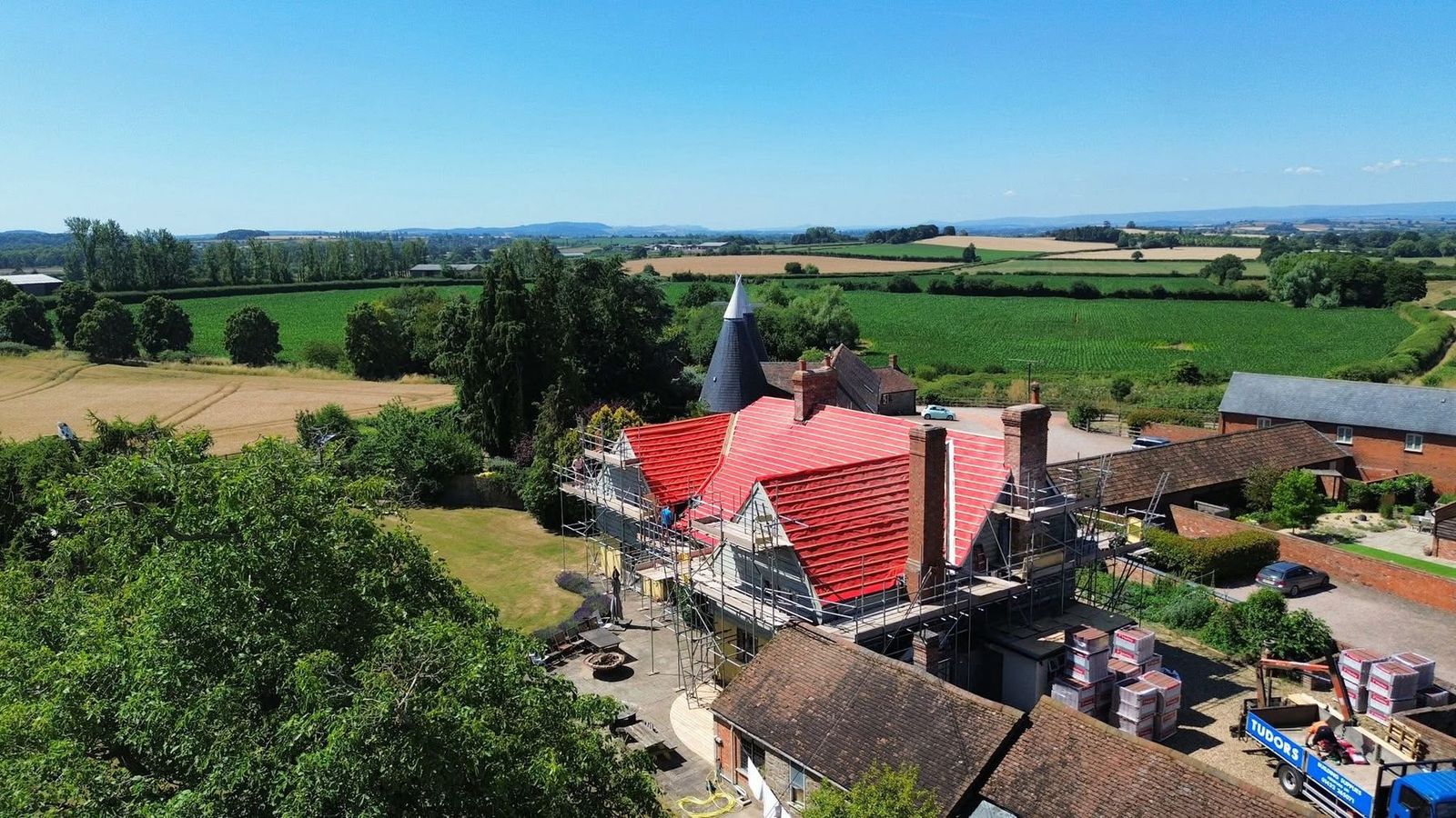 Country house re-roof in progress, the stripped roof covered in breathable underlay and new battens with scaffolding to every elevation and an oast house beyond